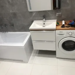 Nowoczesna łazienka z wanną, umywalką z szafką i pralką Samsung, na drewnianym blacie leżą narzędzia i akcesoria glazurnicze, szare płytki marmurowe na ścianie, biała podłoga z połyskiem.