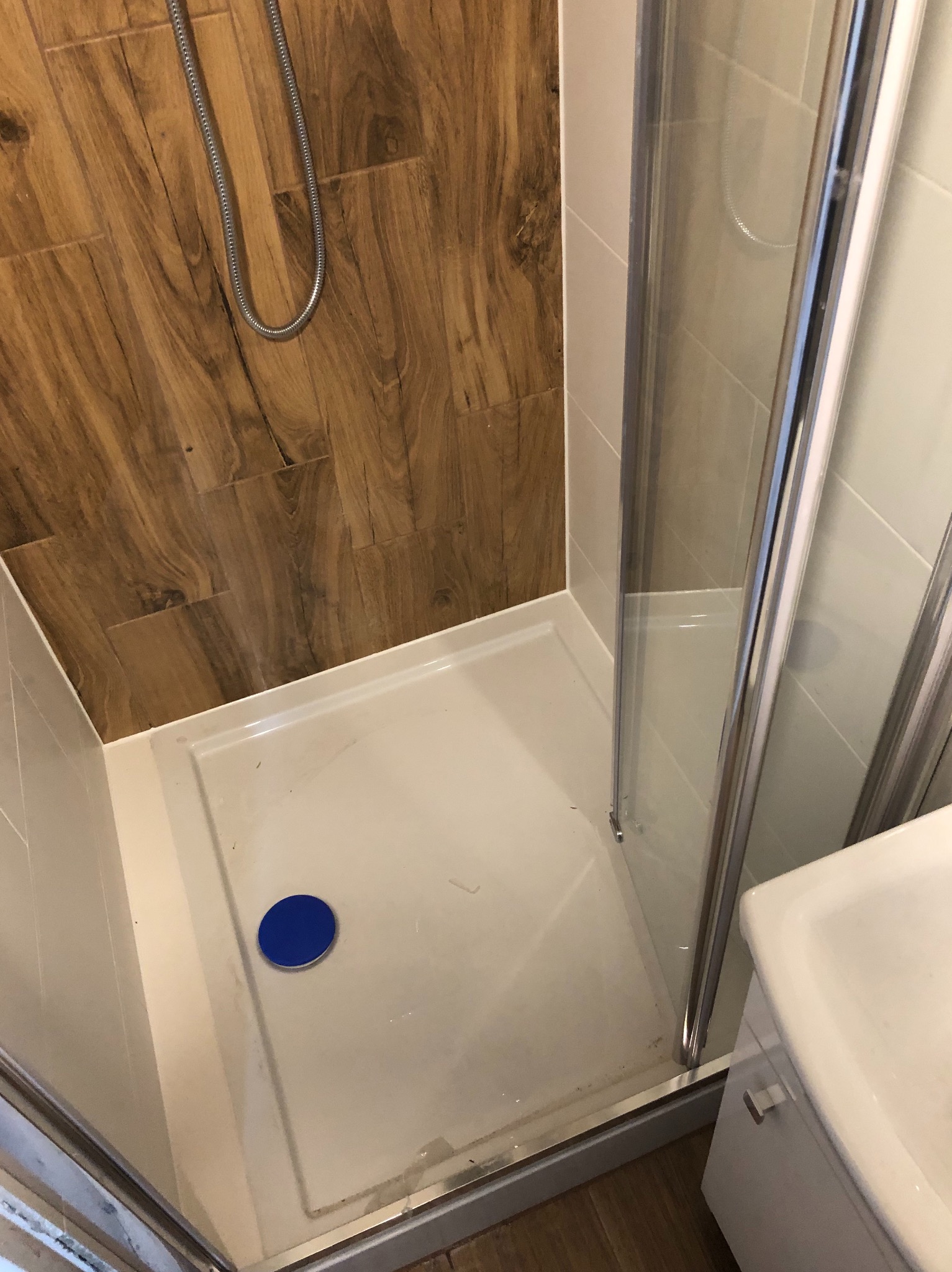 Kabina prysznicowa z brodzikiem i panelami imitującymi drewno na ścianie, widoczny niebieski odpływ i chromowany wąż prysznicowy.