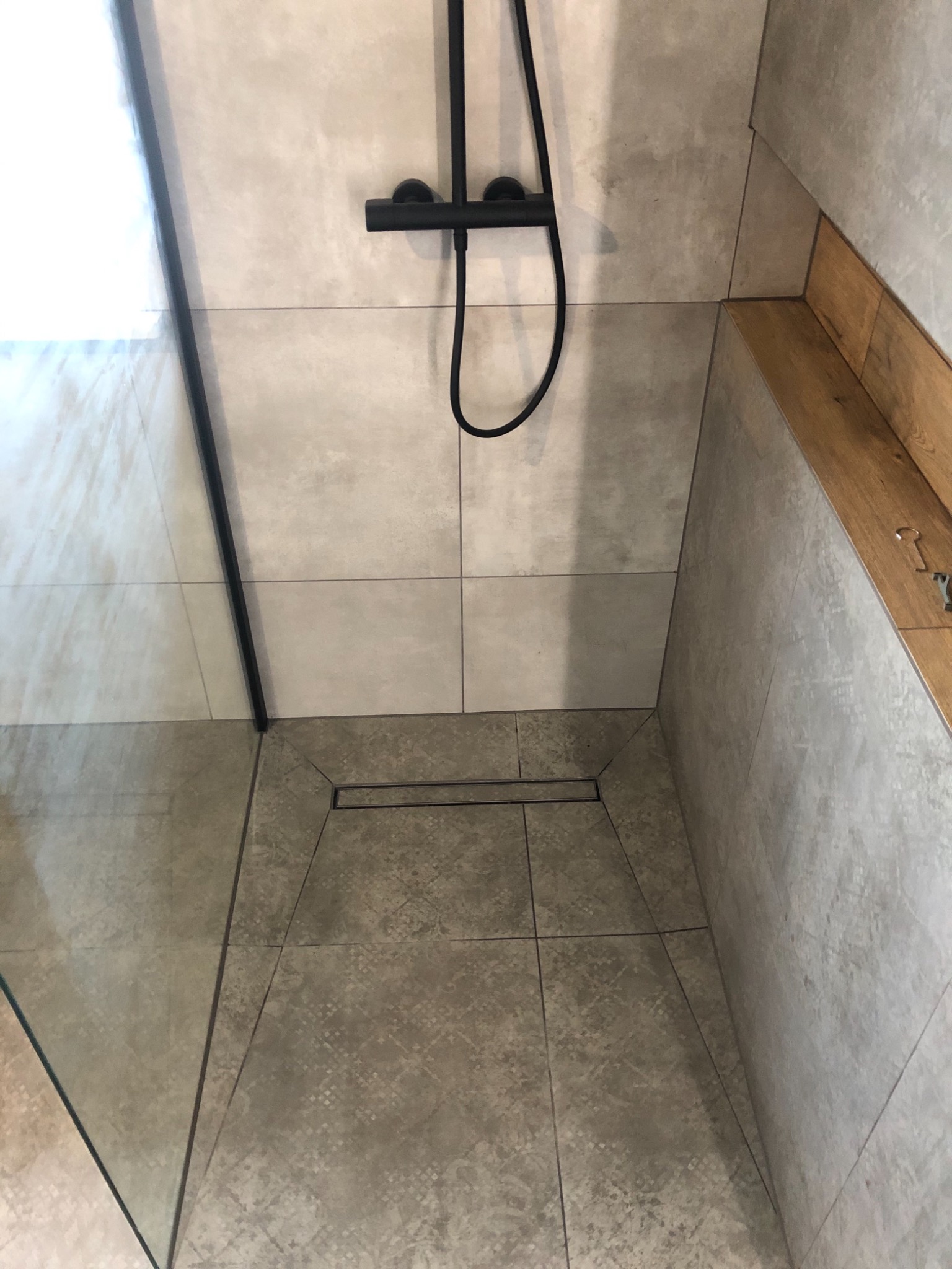 Nowoczesny prysznic walk-in z czarną armaturą, odpływem liniowym i szarą płytką imitującą kamień, wykończony drewnianym blatem.