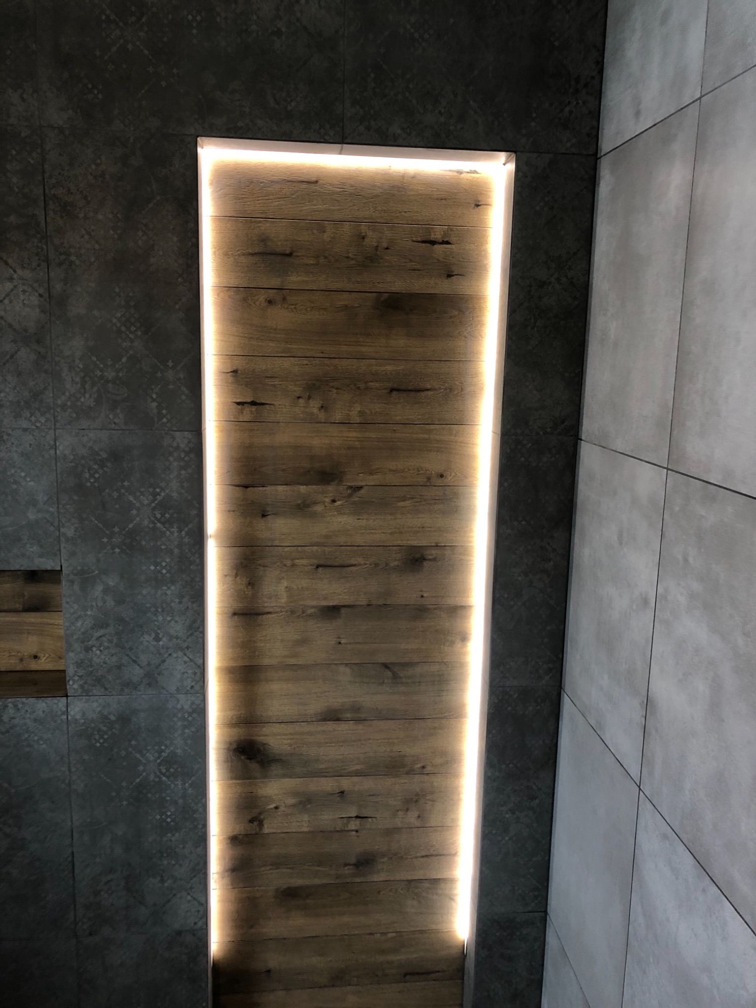 Wnęka w ścianie wyłożona drewnem z oświetleniem LED, sąsiadująca z szarą płytką z wzorem i gładką szarą płytką.