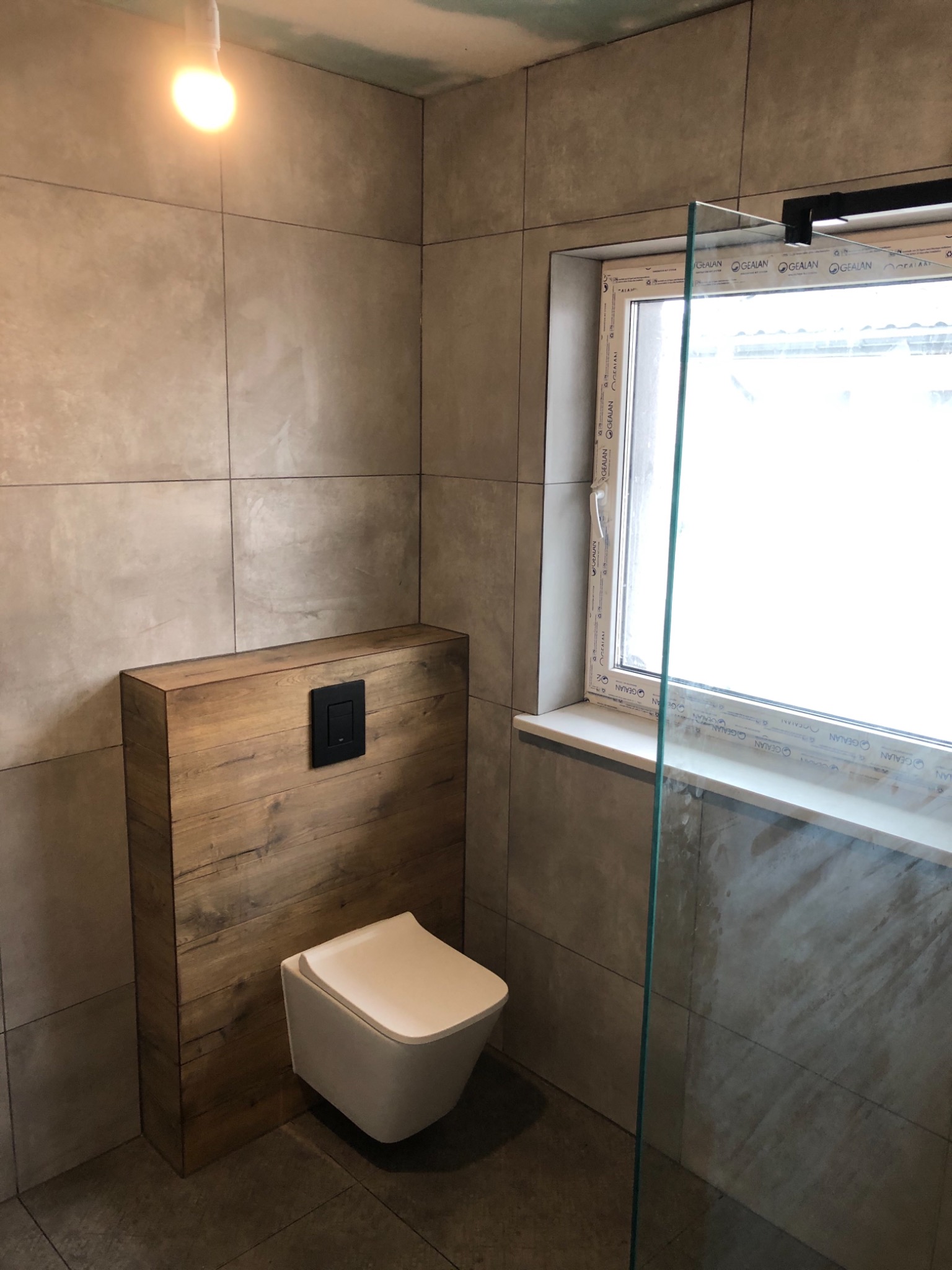 Nowoczesna łazienka z beżowymi płytkami, podwieszana toaleta z drewnianą obudową, szklana kabina prysznicowa i okno PCV z widocznym logotypem producenta.