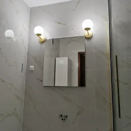 Wykończona łazienka z marmurowymi płytkami, oświetleniem punktowym w stylu retro i kabiną prysznicową z elementami w kolorze złotym.