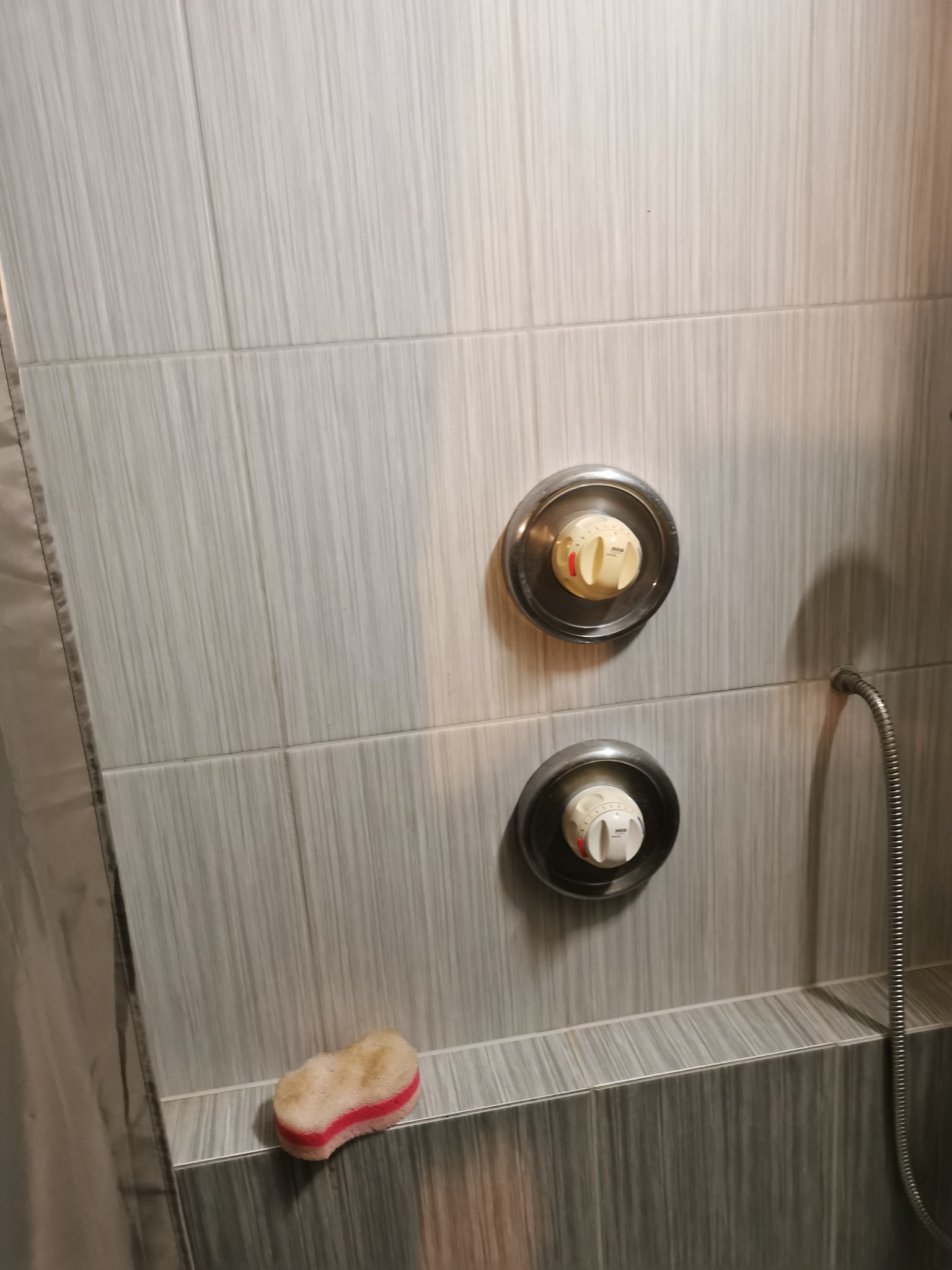Szare płytki w kabinie prysznicowej z dwoma okrągłymi regulatorami temperatury i gąbką do mycia w rogu.