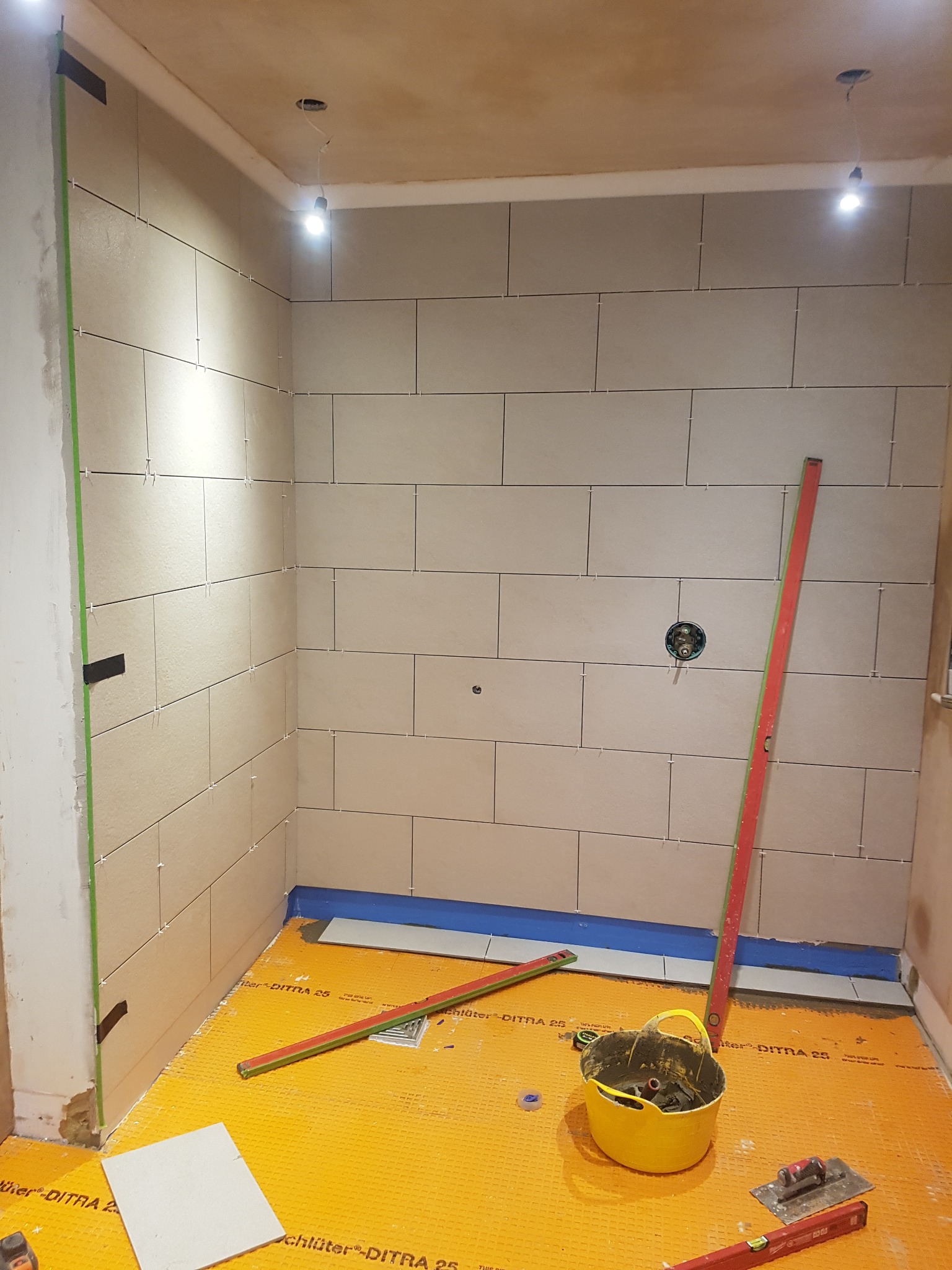 Montaż płytek ściennych w łazience, widoczne poziomice, wiadro z zaprawą i membrana podłogowa DITRA 25.
