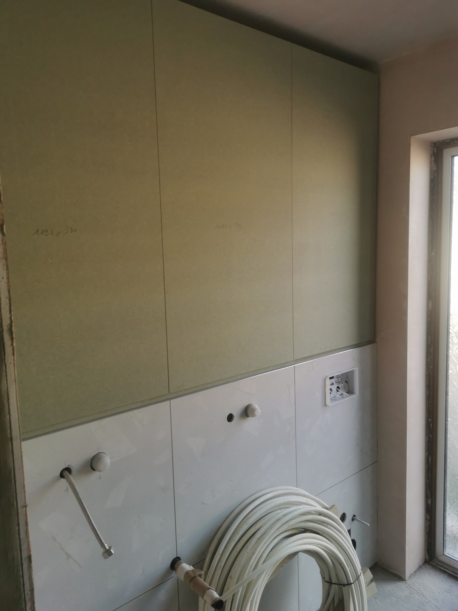 Ściana łazienki w trakcie wykańczania: białe płytki ceramiczne częściowo położone, górna część ściany wyłożona zielonymi płytami gipsowo-kartonowymi z odręcznymi notatkami, widoczne rury...