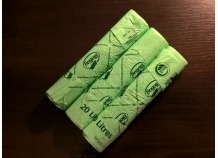 Trzy zwinięte rolki jasnozielonych, kompostowalnych worków na śmieci o pojemności 20 litrów leżące ukośnie na ciemnobrązowym tle. Worki posiadają nadrukowane logo i napisy.