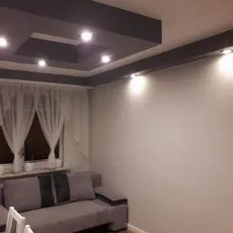 Nowoczesny salon z szarą, podwieszaną konstrukcją sufitową z oświetleniem punktowym, jasnymi ścianami i drewnianą podłogą. W tle widoczny szary narożnik i okno z białymi firankami.