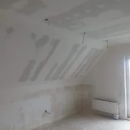 Poddasze w trakcie remontu: ściany i sufit pokryte płytami kartonowo-gipsowymi, widoczne szpachlowanie łączeń, wystające kable elektryczne, grzejnik zamontowany przy ścianie szczytowej.