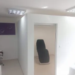 Jasne, minimalistyczne wnętrze gabinetu z białymi ścianami, kremową podłogą z płytek, oświetleniem sufitowym i czarnym fotelem lekarskim w tle, widziane z perspektywy wejścia.