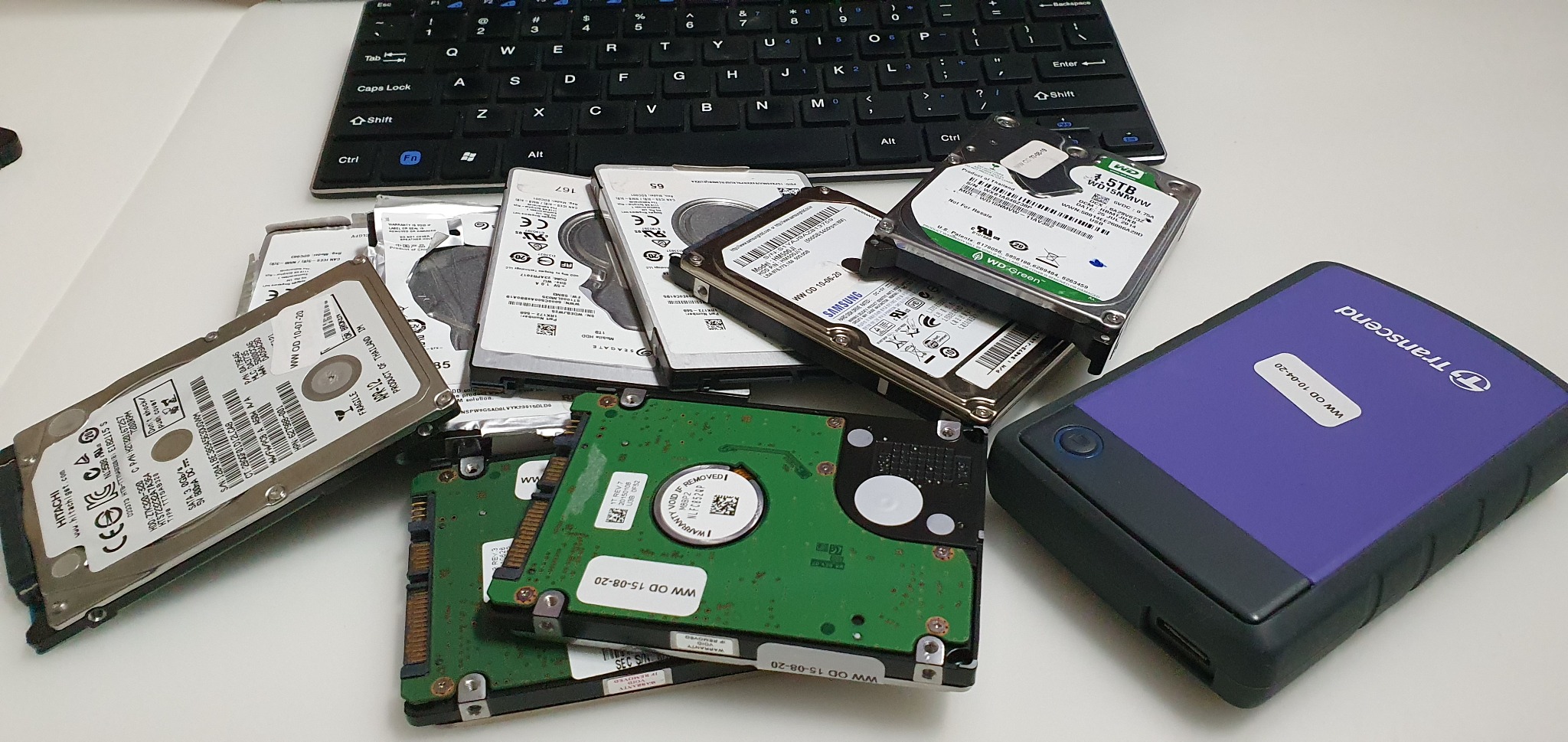 Stos różnorodnych dysków twardych (HDD) różnych producentów i rozmiarów, leżących obok przenośnej obudowy dysku Transcend i klawiatury na białym biurku.