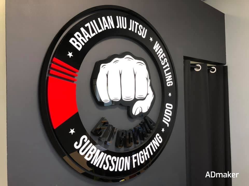 Okrągły, podświetlany szyld z motywem pięści i nazwami sportów walki: Brazilian Jiu Jitsu, Wrestling, Judo, Submission Fighting. Czerwone akcenty i czarne, błyszczące litery na szarym tle ściany.