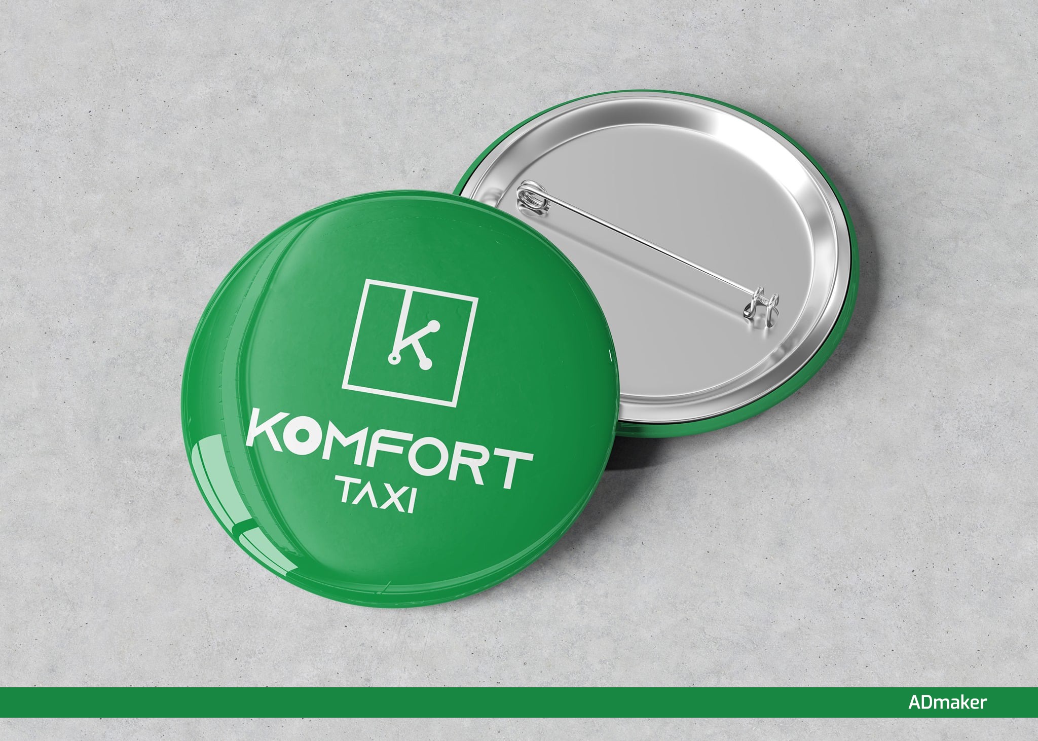 Dwie okrągłe przypinki z logo 'Komfort Taxi' w kolorze zielonym i białym, leżące na szarym tle z widocznym mechanizmem mocowania.