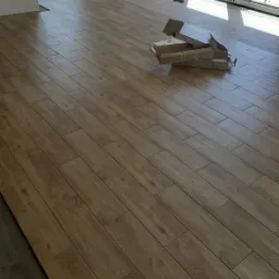 Ułożona podłoga z płytek imitujących drewno w jasnym odcieniu brązu, z otwartymi kartonami po płytkach w rogu pomieszczenia, widoczne okno z widokiem na ogród.