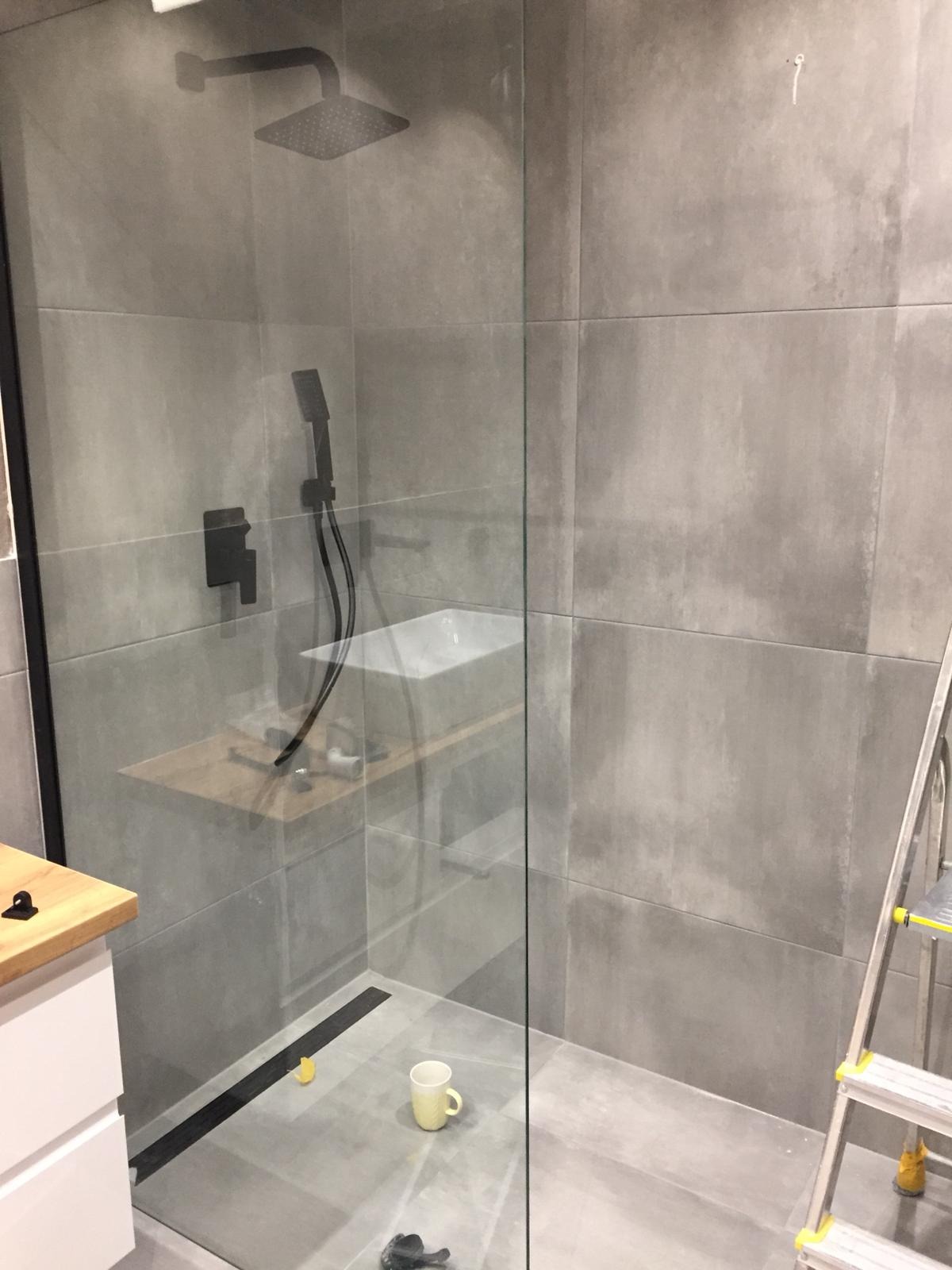 Szara łazienka z kabiną prysznicową typu walk-in, czarną armaturą, odpływem liniowym i kubkiem na podłodze, widoczna drabina malarska.