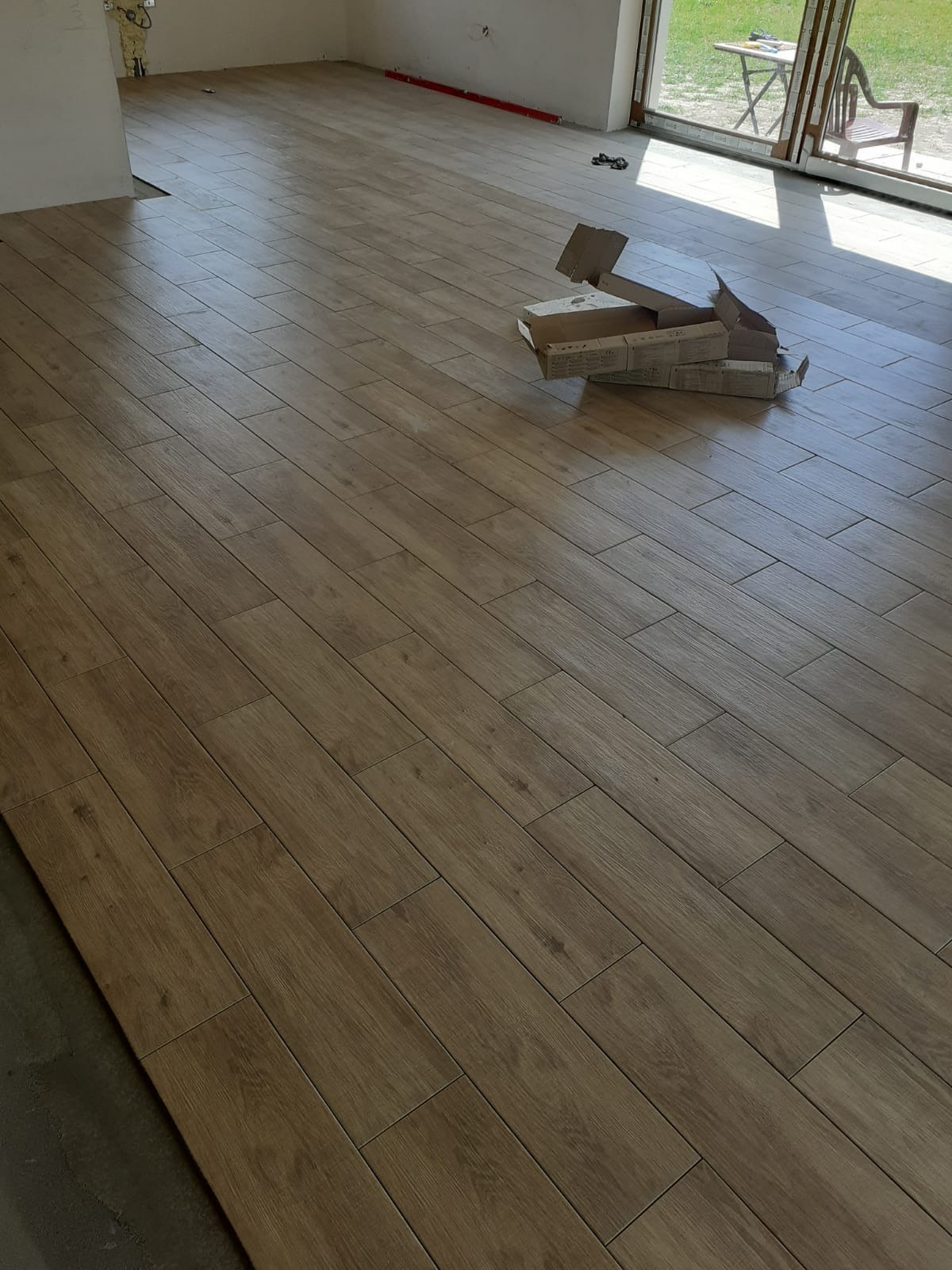 Ułożona podłoga z płytek imitujących drewno w jasnym odcieniu brązu, z otwartymi kartonami po płytkach w rogu pomieszczenia, widoczne okno z widokiem na ogród.