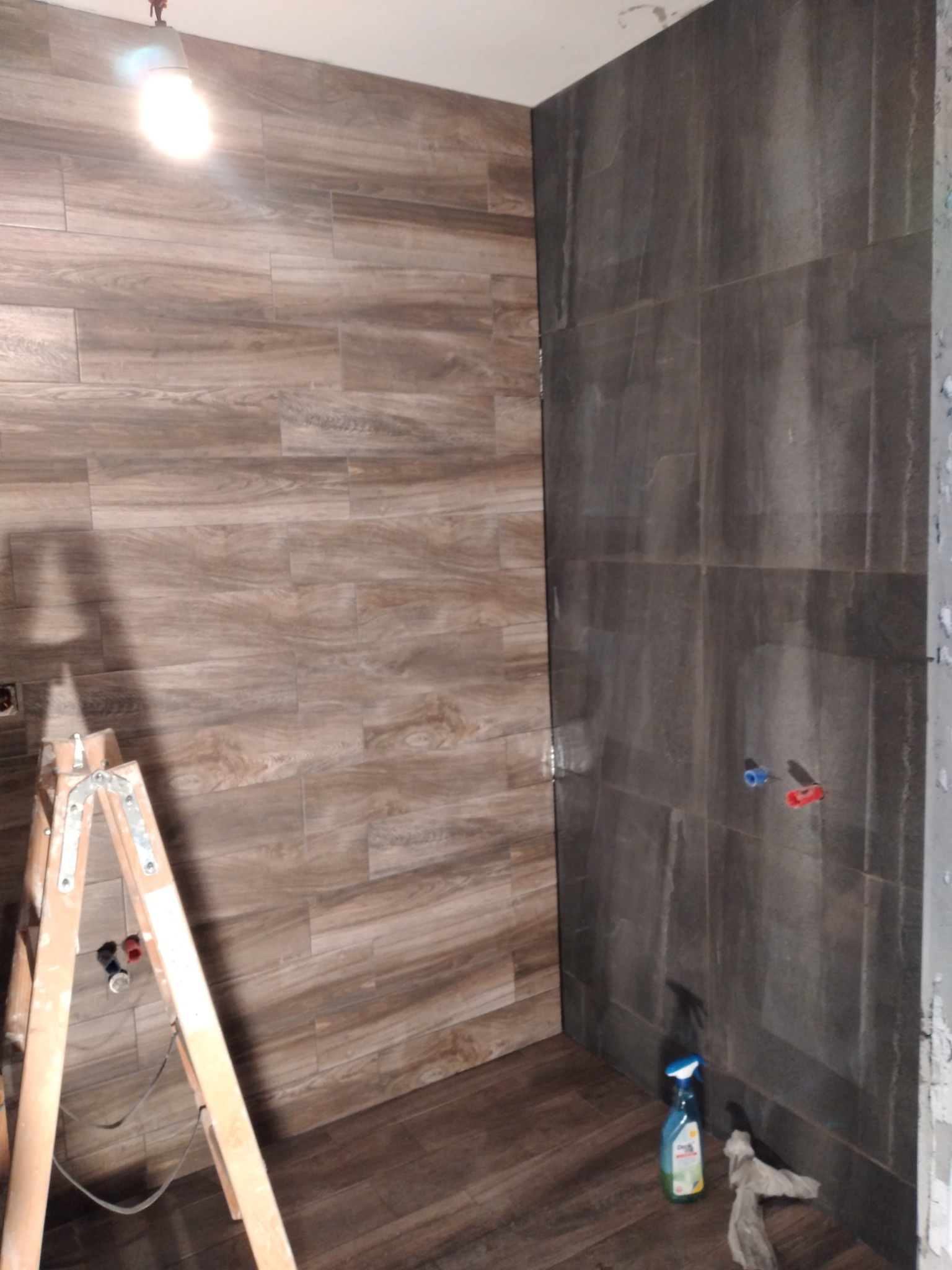 Układanie płytek imitujących drewno na ścianie w łazience, widoczna drabina malarska i chemia czyszcząca.