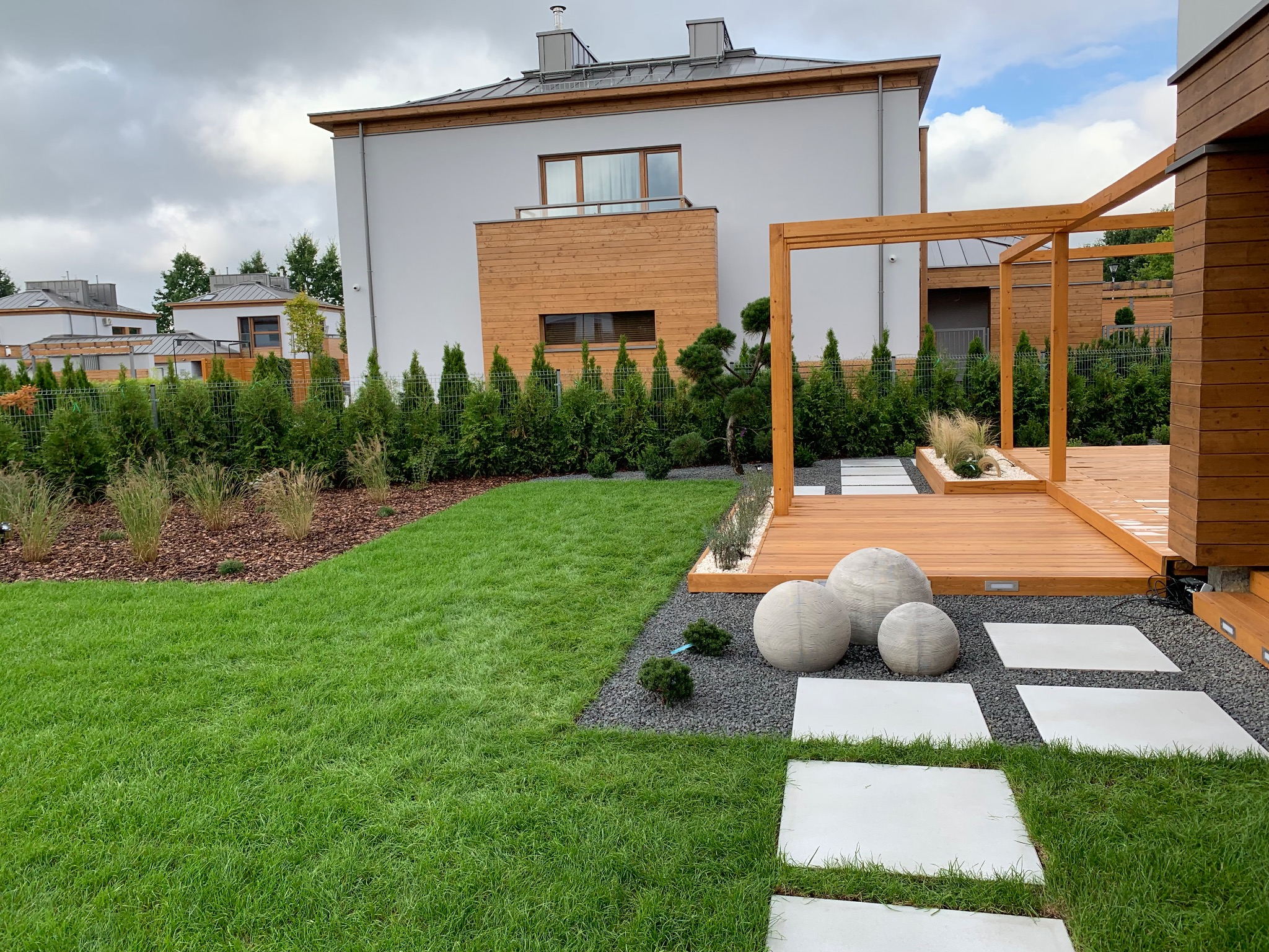 Nowoczesny ogród z drewnianym tarasem i pergolą, ścieżką z betonowych płyt, kulistymi dekoracjami i równo przystrzyżonym trawnikiem na tle domu.