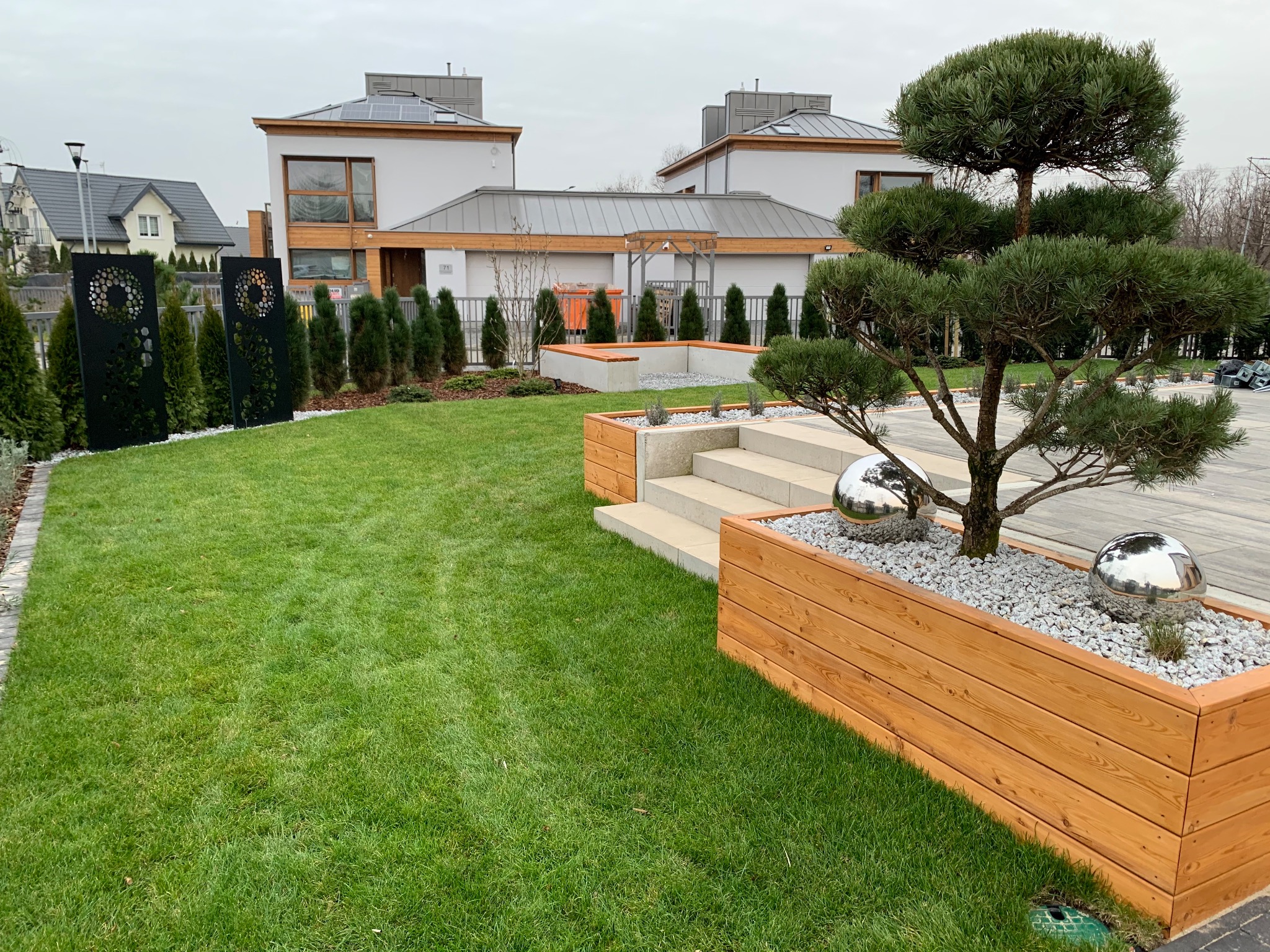 Nowoczesny ogród z równo przystrzyżonym trawnikiem, ozdobnymi panelami, formowanymi drzewami w drewnianych donicach wypełnionych białym żwirem i metalowymi kulami, oraz schodami z jasnego kamienia.