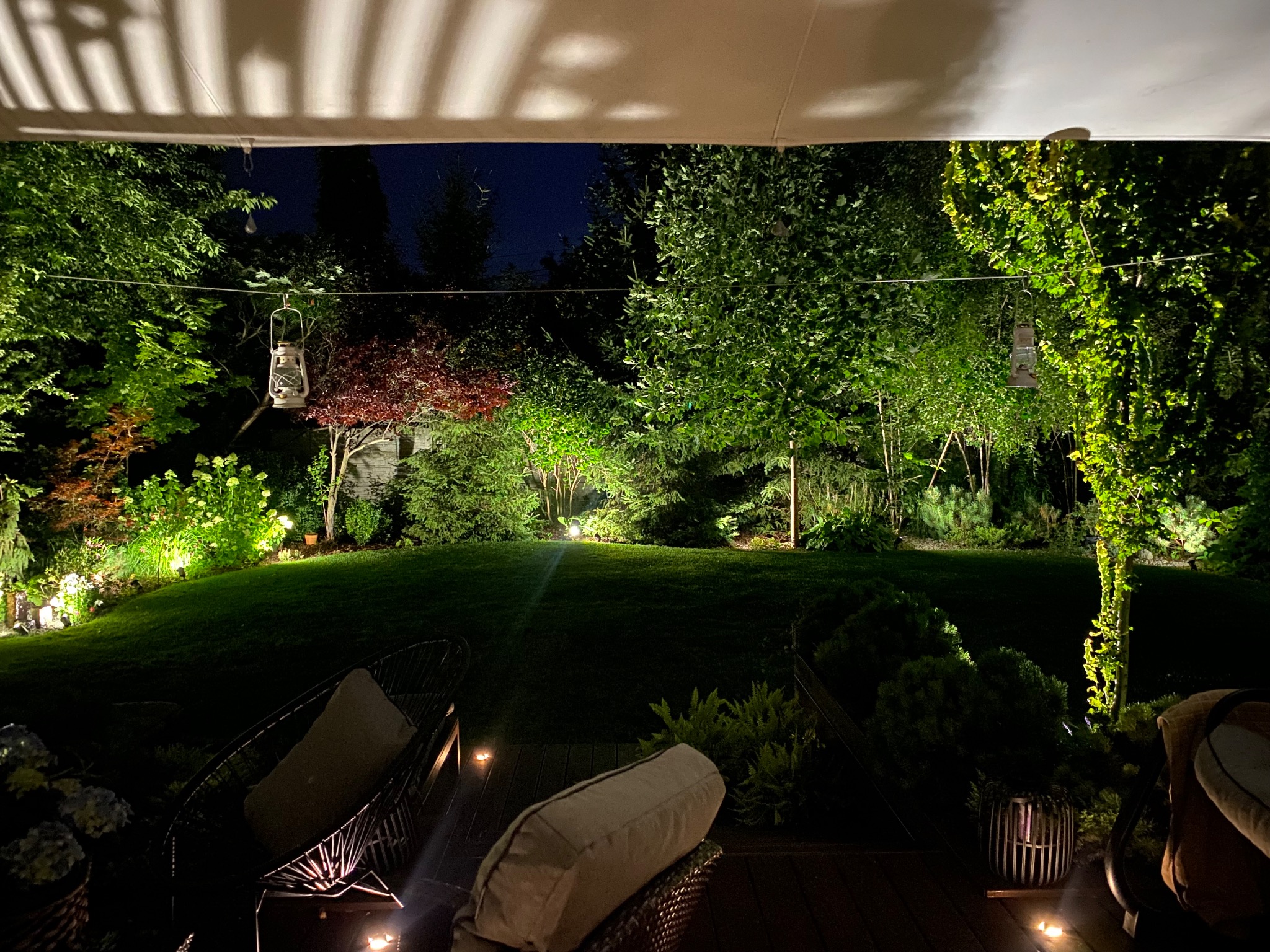 Oświetlony ogród nocą z widokiem z tarasu, wiszące lampiony nad trawnikiem, podświetlone drzewa i krzewy, meble ogrodowe na pierwszym planie.