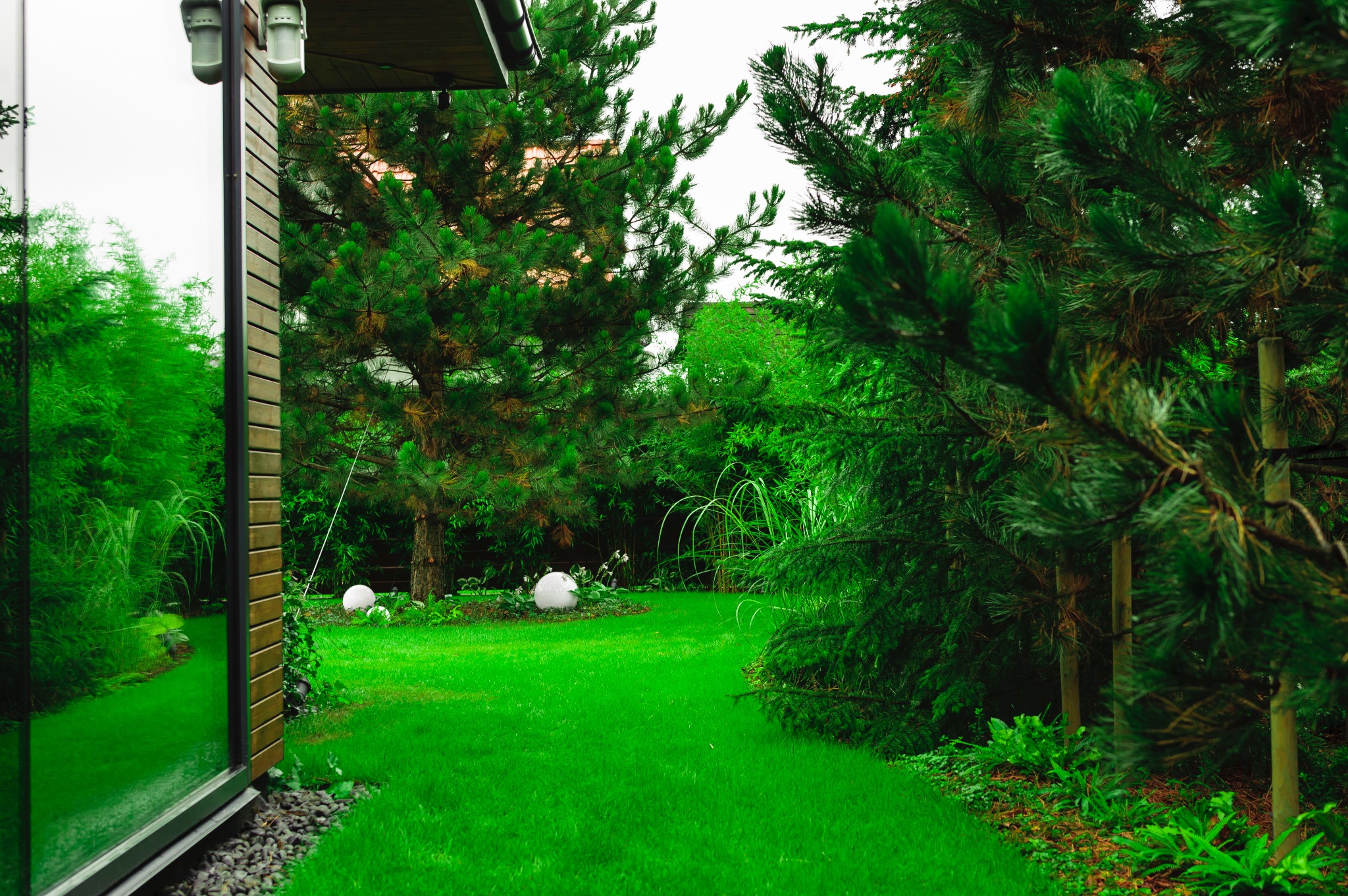 Zadbany trawnik z ozdobnymi kulami oświetleniowymi w ogrodzie, widok zza nowoczesnego budynku z dużymi oknami.