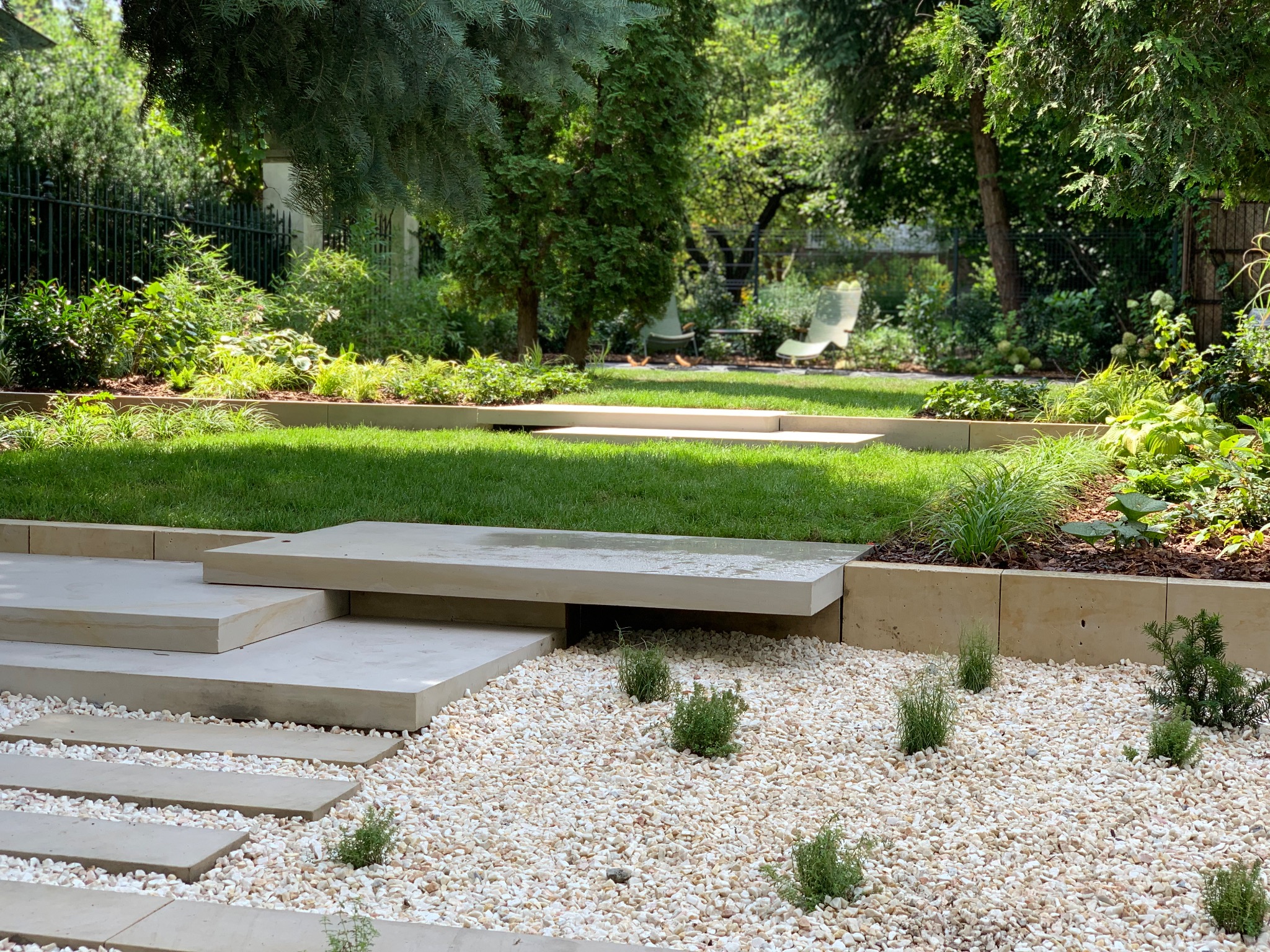 Nowoczesny ogród z trawnikiem, kamienną ścieżką i podwyższonymi rabatami, wykończony jasnym żwirem.