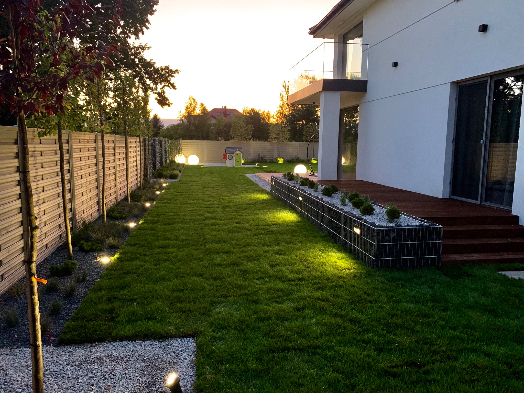 Ogród z równo przystrzyżonym trawnikiem, oświetlony kulistymi lampami i lampami wzdłuż ogrodzenia, z nowoczesnym domem w tle podczas zachodu słońca.