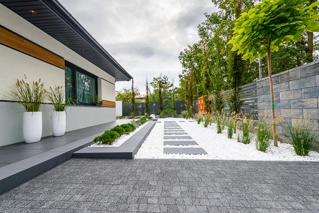 Nowoczesny ogród z szarą kostką brukową, białymi kamykami, ścieżką z płyt i zielenią, widziany z perspektywy podjazdu.