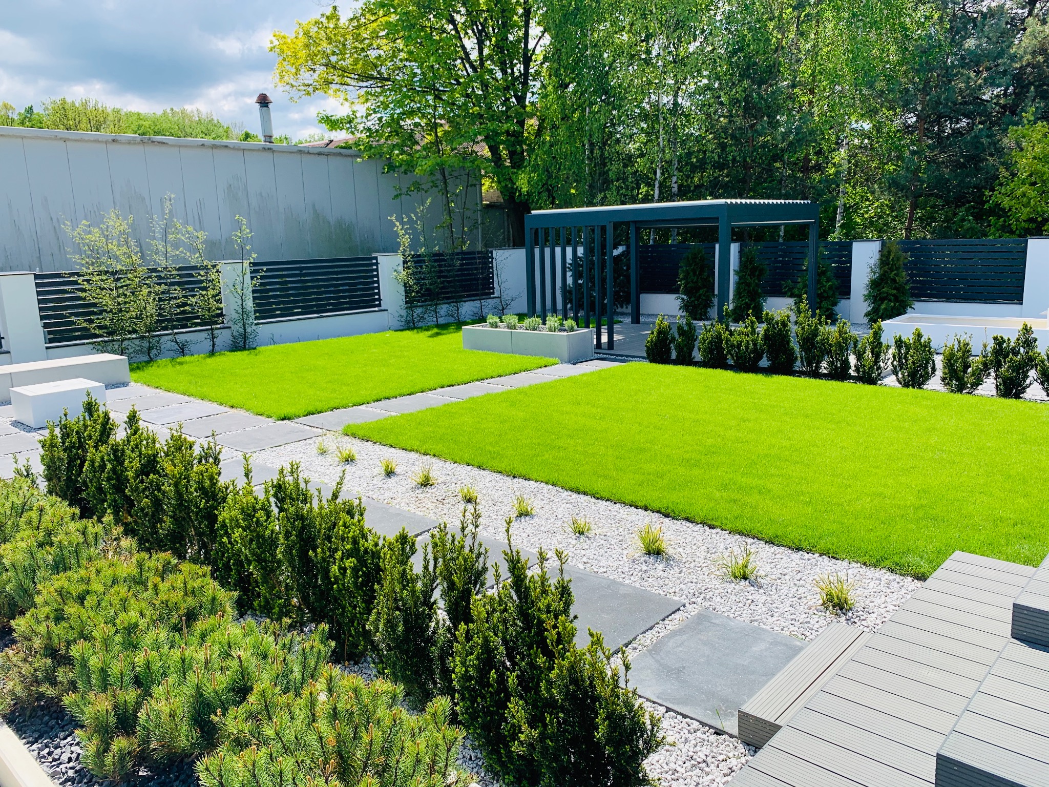Perfekcyjnie utrzymany ogród z równo przystrzyżonym trawnikiem, ścieżką z płyt granitowych i białego żwiru, niskimi krzewami iglastymi oraz nowoczesną pergolą w tle.