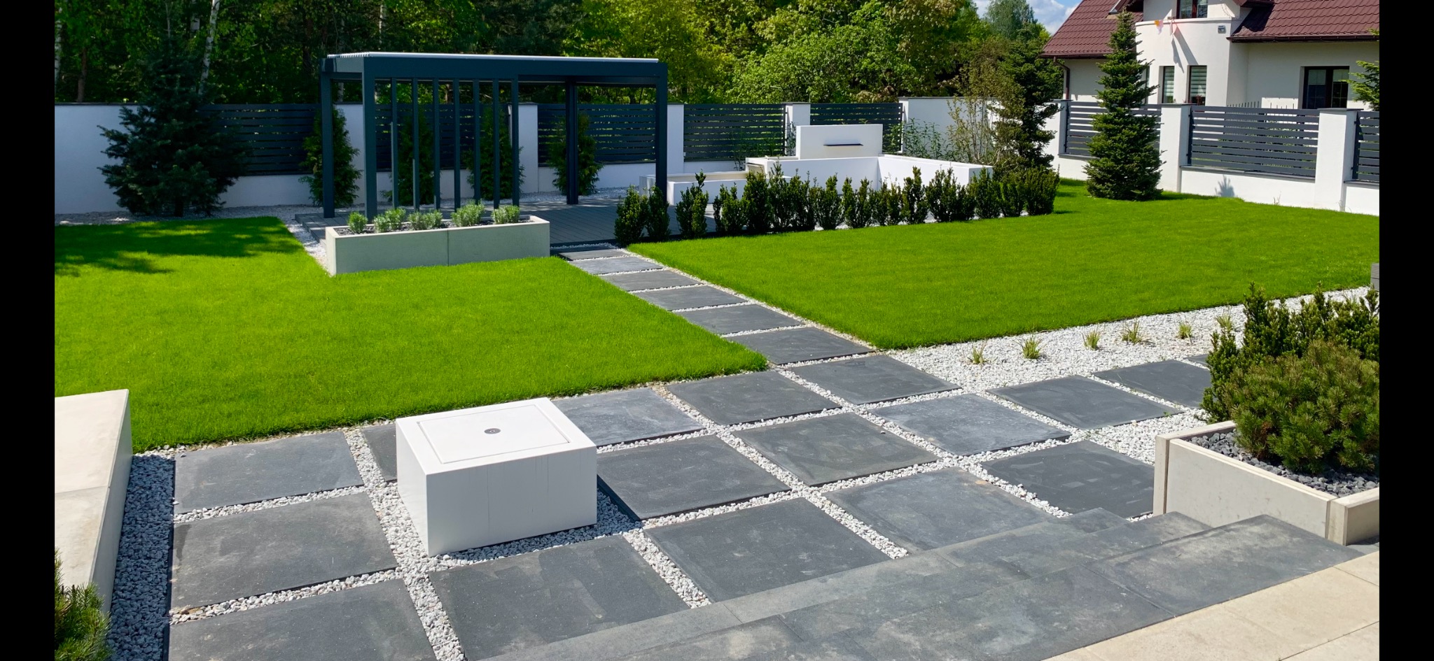 Nowoczesny ogród z równo przystrzyżonym trawnikiem, ścieżką z kwadratowych płyt oddzielonych białym żwirem, minimalistyczną pergolą i białym elementem wodnym w tle.