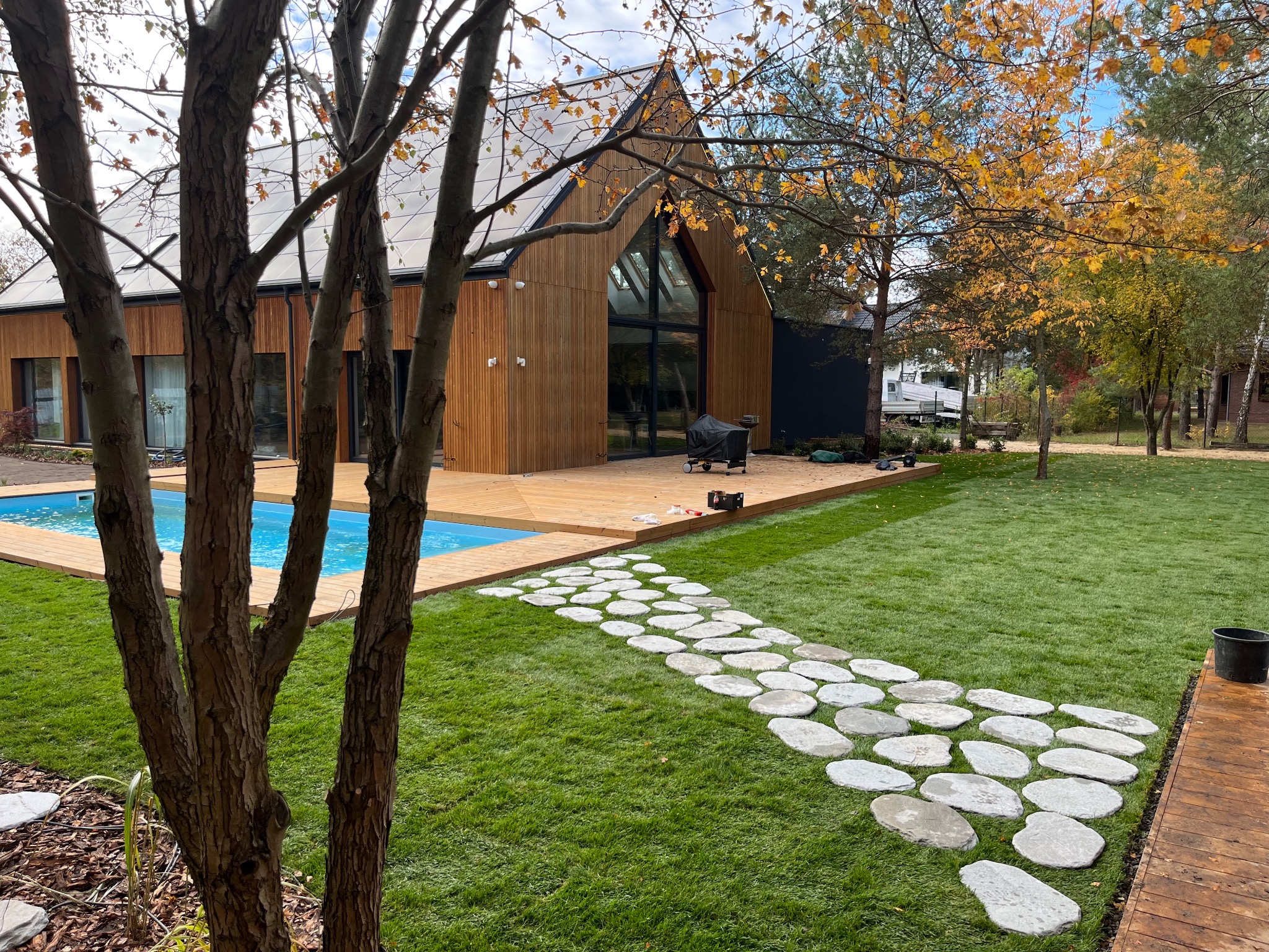 Nowoczesny ogród z basenem i ścieżką z kamieni prowadzącą przez trawnik. Drewniany taras przylega do domu z elewacją z jasnego drewna. Jesienne drzewo w tle.