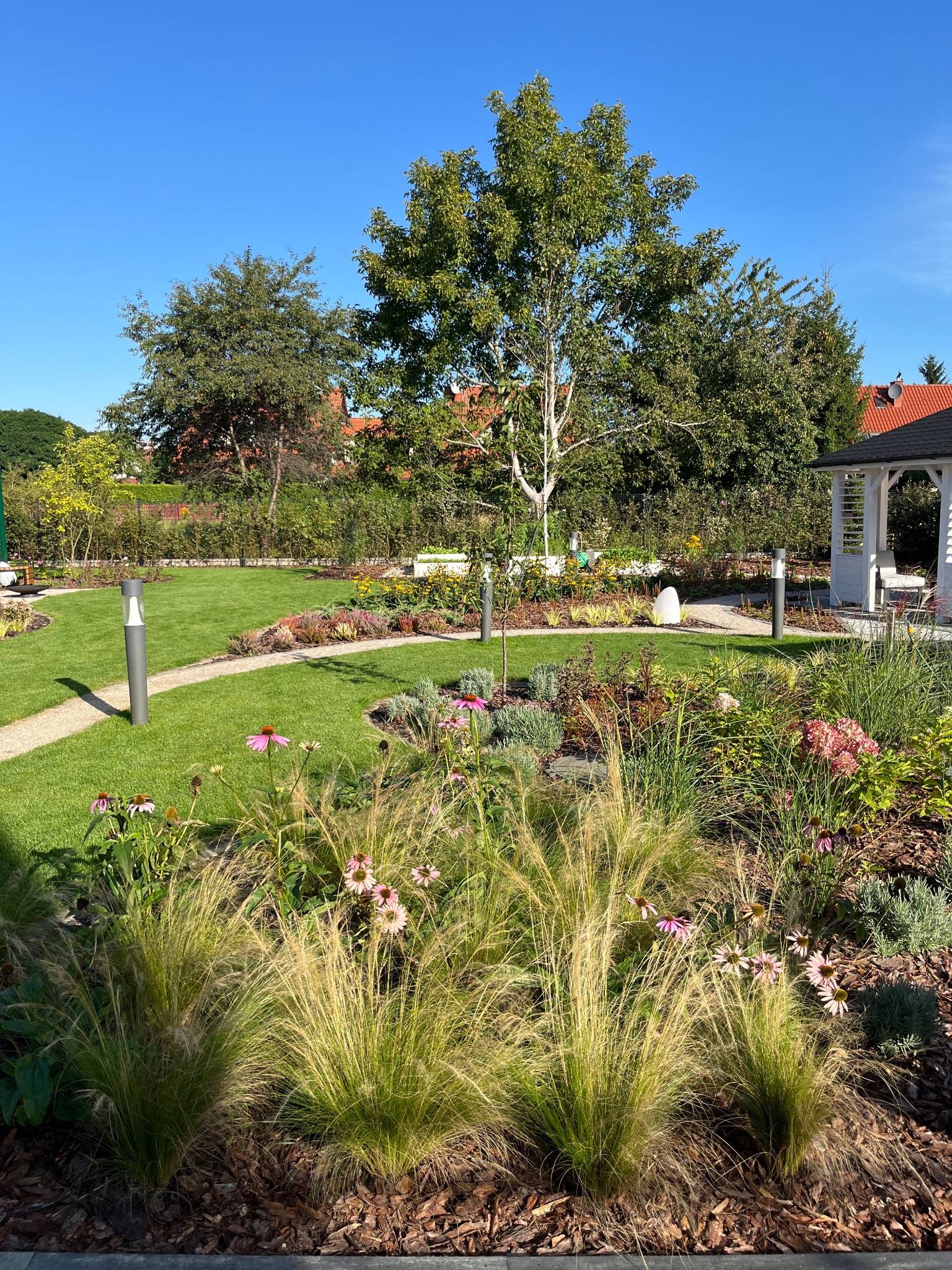 Zadbany ogród z trawnikiem, ścieżką z kamienia, rabatami kwiatowymi i ozdobnymi trawami. Widoczna altana i oświetlenie ogrodowe. Słoneczny dzień.