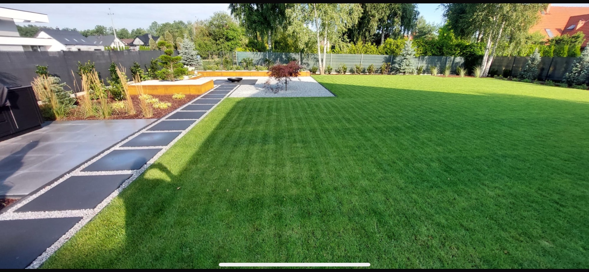 Nowoczesny ogród z równo przystrzyżonym trawnikiem, ścieżką z ciemnych płyt i rabatami z ozdobnymi roślinami. Minimalistyczny design z elementami dekoracyjnymi.