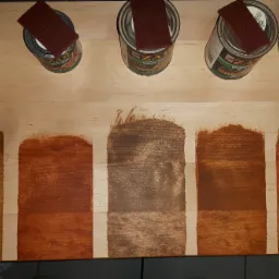 Próbki różnych odcieni bejcy na drewnie, z puszkami bejcy ZAR i gąbkami aplikacyjnymi na górze, prezentujące efekt kolorystyczny po nałożeniu na jasne drewno.