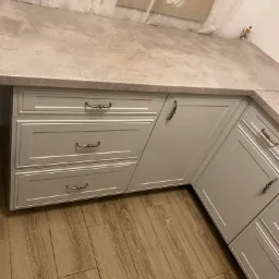 Fragment zabudowy kuchennej z szarymi frontami szafek i szuflad z ozdobnymi uchwytami, widoczne także drewnopodobne płytki podłogowe i fragment blatu przy oknie z firanką.