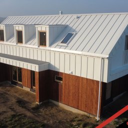 PHU Optimum Dachy Jaszczerski Marcin - Nowoczesny dom z białym dachem z blachy na rąbek stojący i drewnianą elewacją, widok z góry, w tle pola i zabudowa.