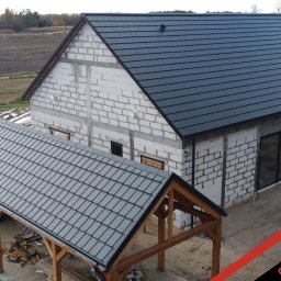 PHU Optimum Dachy Jaszczerski Marcin - Nowoczesny dom w trakcie budowy z ciemnoszarą dachówką ceramiczną i zadaszeniem tarasu o identycznym pokryciu. Widoczny surowy stan ścian z betonu komórkowego.