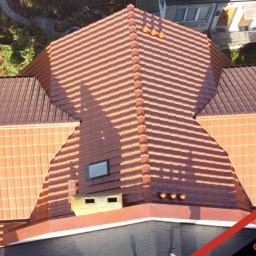 PHU Optimum Dachy Jaszczerski Marcin - Widok z góry na nowy, pomarańczowy dach z dachówki ceramicznej z oknem dachowym i kominem, otoczony zielenią. Widoczne detale wykończenia i elementy wentylacyjne.
