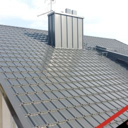 PHU Optimum Dachy Jaszczerski Marcin - Szary dach z dachówki modułowej, komin z blachy na tle błękitnego nieba. Widok z góry ukazuje regularny wzór pokrycia i detale wykończenia.