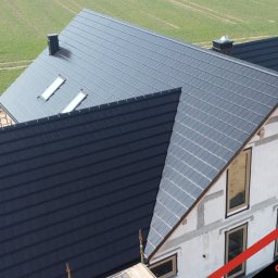 PHU Optimum Dachy Jaszczerski Marcin - Nowoczesny dom w trakcie budowy z ciemnoszarą dachówką ceramiczną, widok z góry, okna dachowe, częściowo otynkowana elewacja, zielone pole w tle.