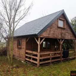 Drewniany domek z szarą blachodachówką na zewnątrz, widoczny z perspektywy ogrodu w pochmurny dzień.