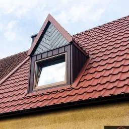 Czerwona dachówka ceramiczna z oknem dachowym i drewnianą konstrukcją szczytu, widok z dołu na tle błękitnego nieba.