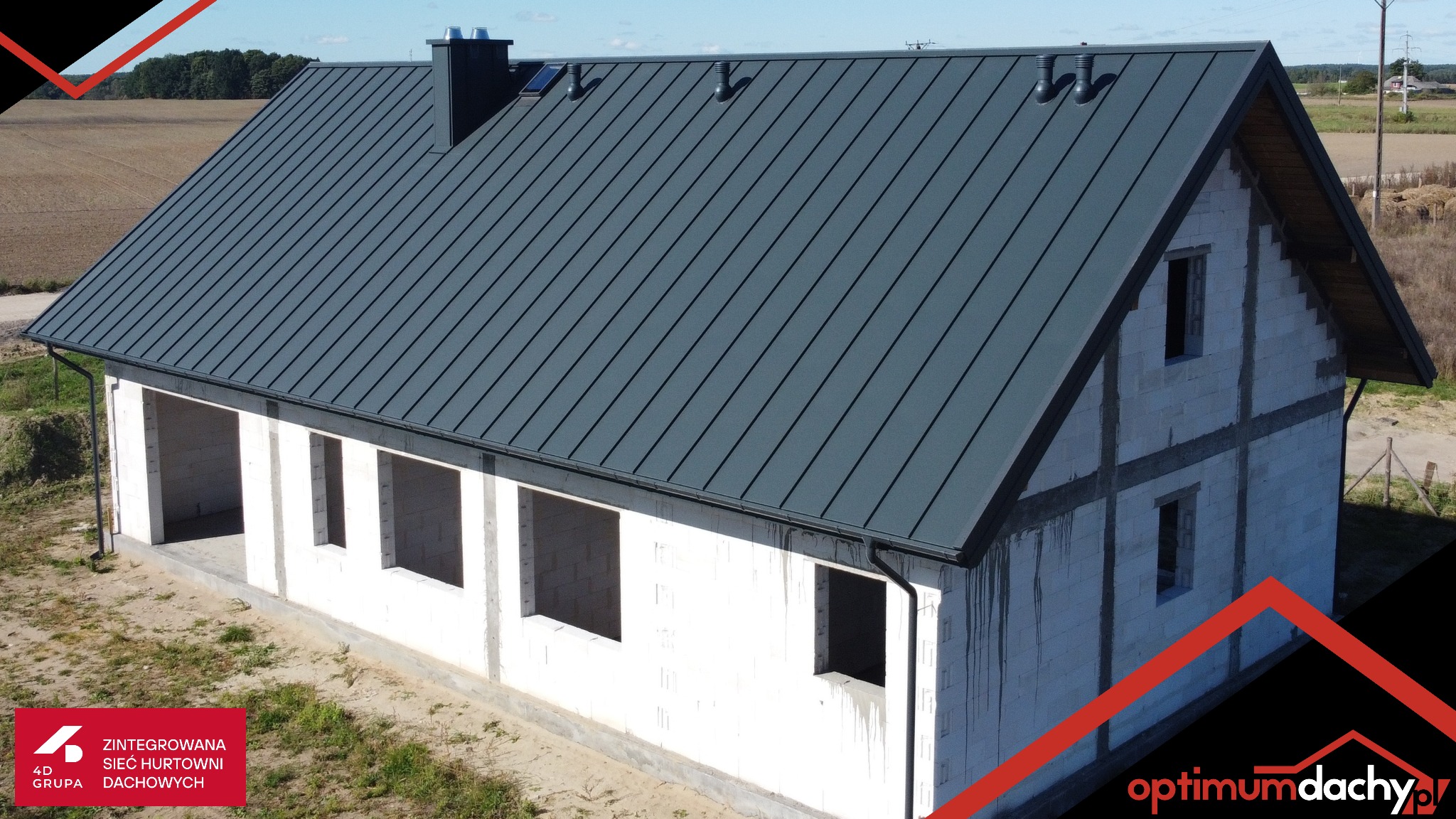 Dom w trakcie budowy z ciemnoszarym dachem z blachy na rąbek stojący, widoczne rynny i komin. Budynek w stanie surowym otwartym.