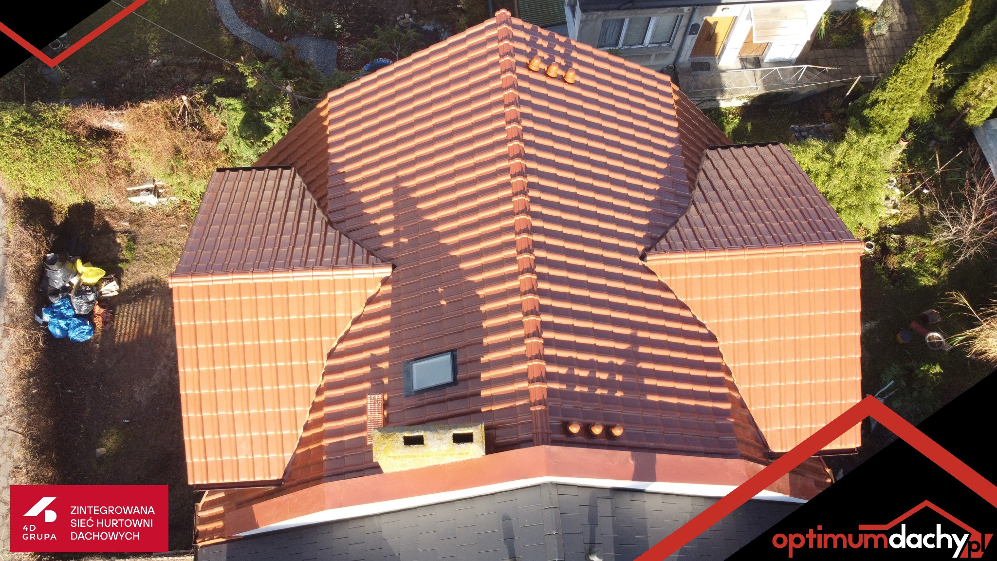 Widok z góry na nowy, pomarańczowy dach z dachówki ceramicznej z oknem dachowym i kominem, otoczony zielenią. Widoczne detale wykończenia i elementy wentylacyjne.