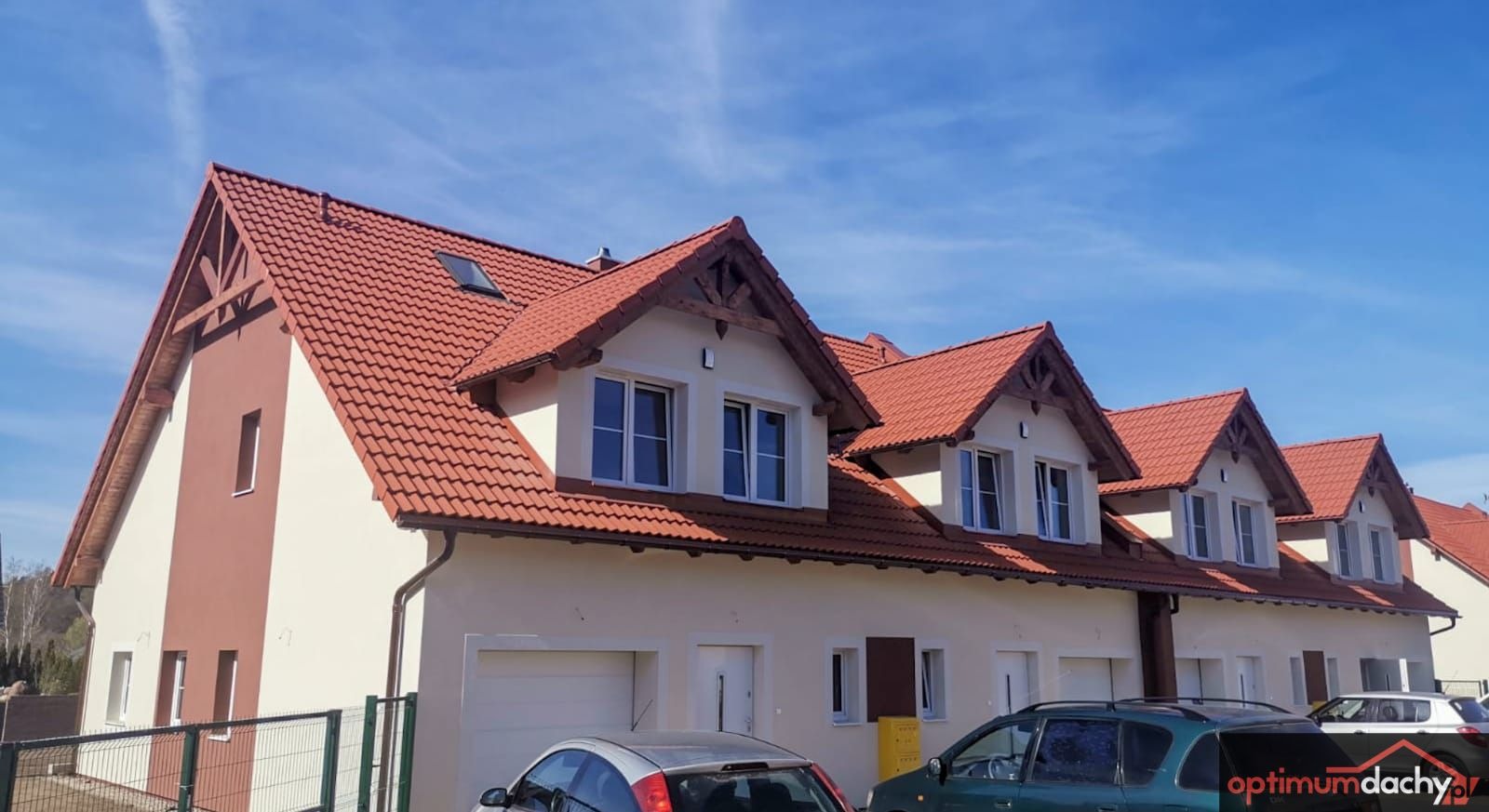 Nowy dach z czerwonej dachówki ceramicznej na szeregu domów z lukarnami i drewnianymi elementami dekoracyjnymi, błękitne niebo w tle.