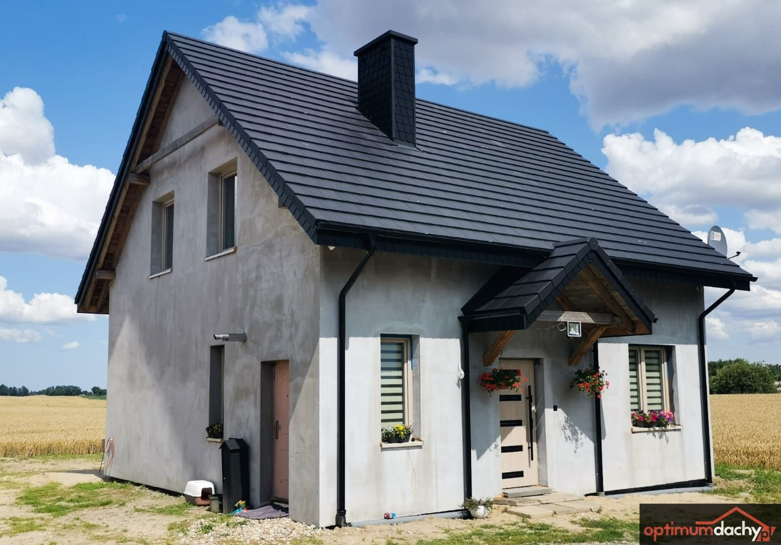 Dom jednorodzinny z ciemną dachówką, widoczne rynny w kolorze czarnym i wykończony komin, elewacja w stanie surowym, w tle pole zboża i błękitne niebo.