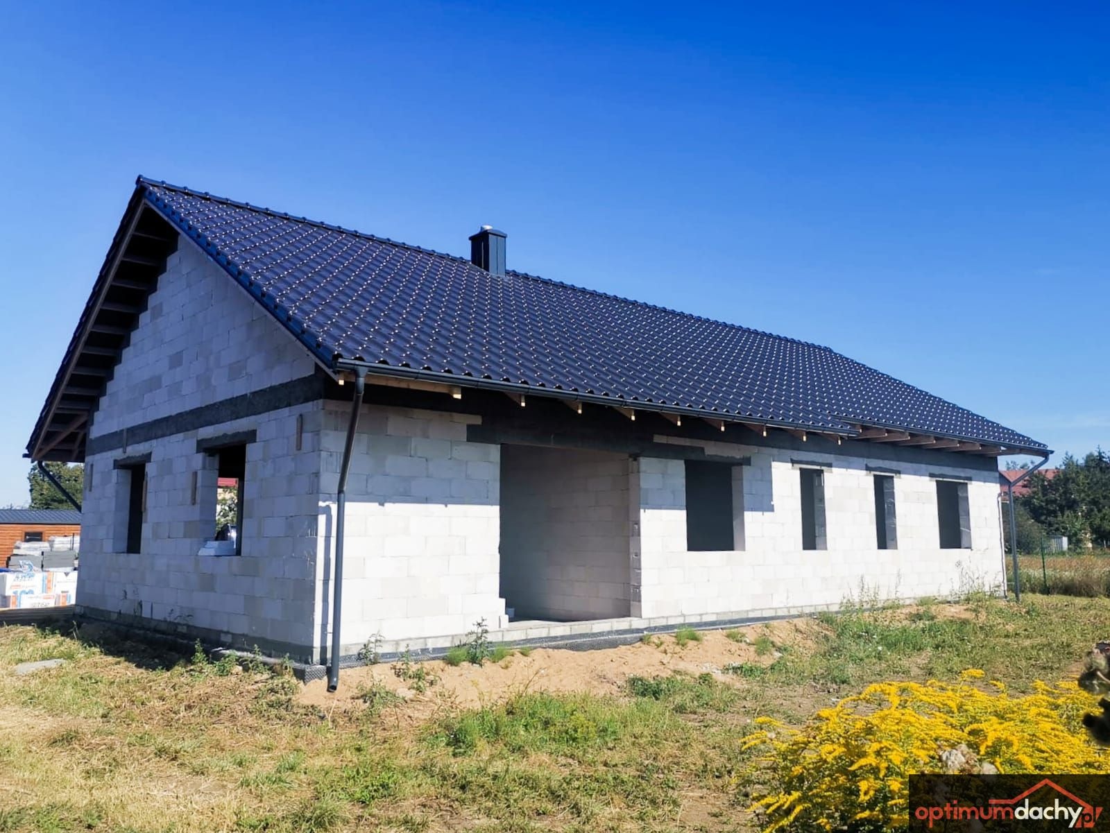 Niedokończony dom jednorodzinny z ciemną dachówką i zamontowanymi rynnami, widoczne bloczki konstrukcyjne i niezabudowane otwory okienne, żółte kwiaty na pierwszym planie.