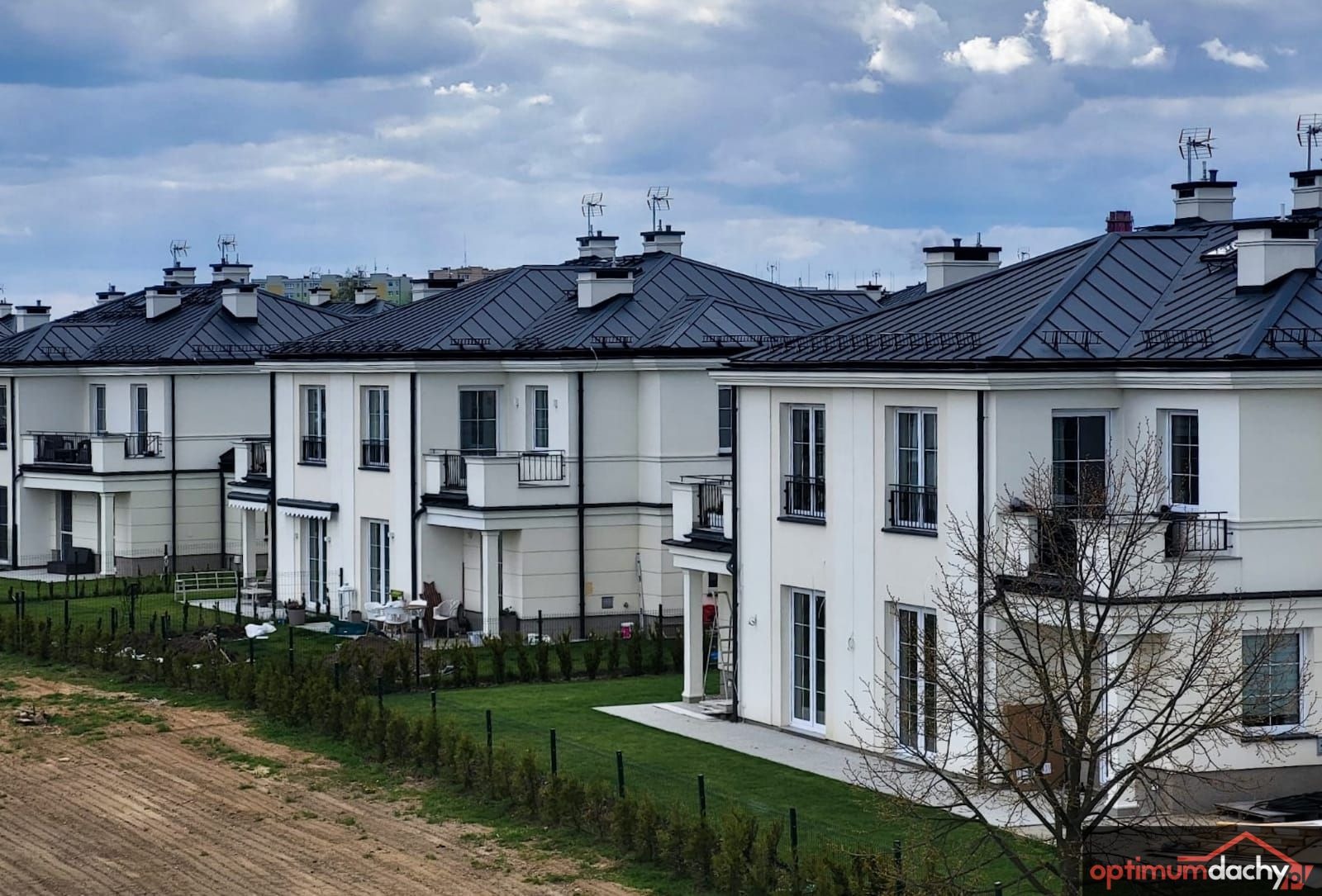 Szereg identycznych, nowoczesnych domów jednorodzinnych w Tczewie z ciemnymi, metalowymi pokryciami. Widok z lekkiego wzniesienia na dachy budynków i zadbane trawniki.