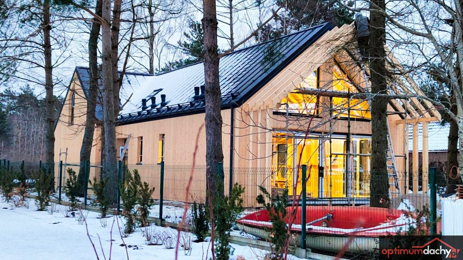 Dom w trakcie budowy z częściowo ukończonym czarnym dachem z blachy, widoczna więźba dachowa i rusztowanie, zimowy krajobraz w tle.