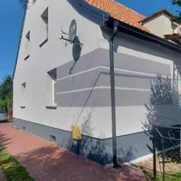 Odnowiona elewacja domu jednorodzinnego w odcieniach szarości z dekoracyjnymi pasami, widoczna antena satelitarna i brukowa ścieżka.