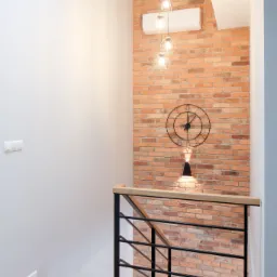 Korytarz ze schodami z drewnianą podłogą i ceglaną ścianą z zegarem, oświetlony wiszącymi lampami i kinkietem, z klimatyzatorem na górze.